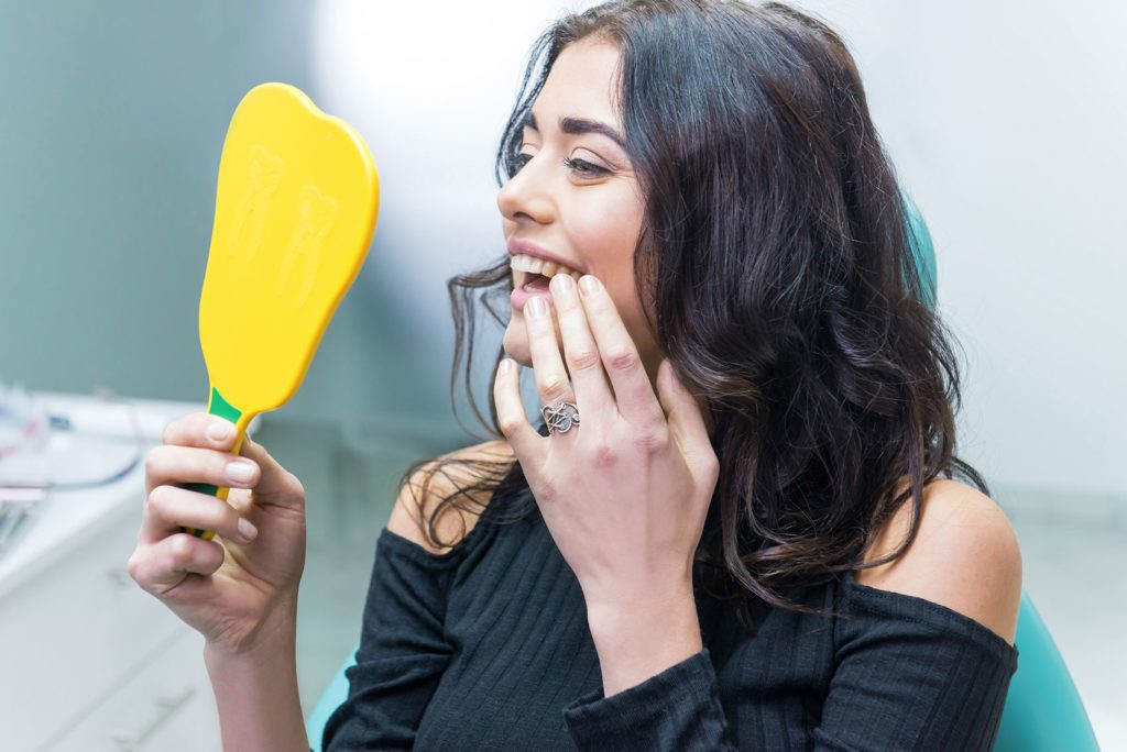 Woman with dark hair examining her teeth in a yellow hand-held mirror